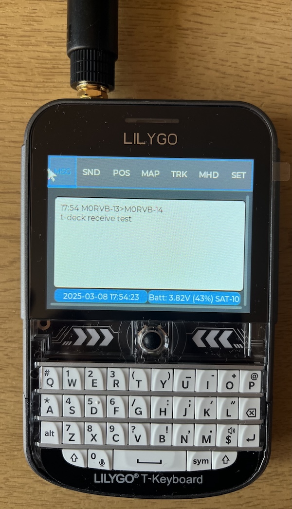 Photo of a Lilygo T-Deck Plus handheld MeshCom terminal showing a white keyboard with black letters and a test message on the LCD screen