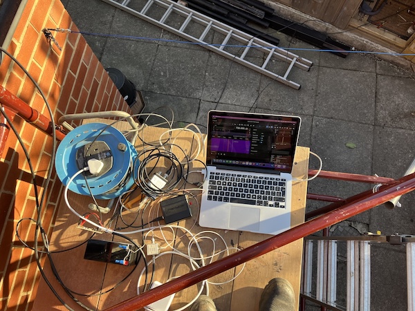 A photo of equipment set on scaffolding planks. There is a MacBook, a bias tee, a GPSDO and various cables.