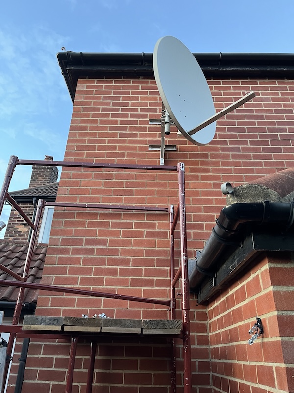 A photo of red scaffolding at the side of a brick house wall and a grey satellite dish mounted on a TK bracket and short section of mast.