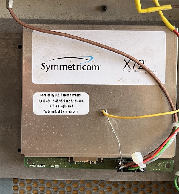 A Symmetricom X72 rubidium oscillator, silver in colour with a white label showing the company logo and another showing patent details.