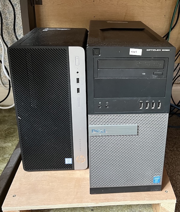A photo of two PCs, on the left an HP and on the right a Dell, sitting on a plywood shelf on the floor.