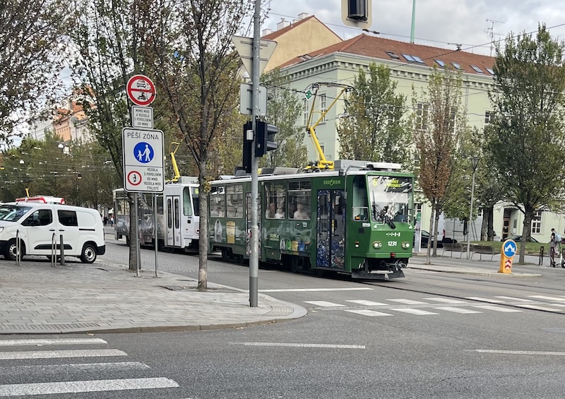 Two joined tram cars, green in front and white behind set against various street furniture. There is a white van to the left, a sign and various trees.