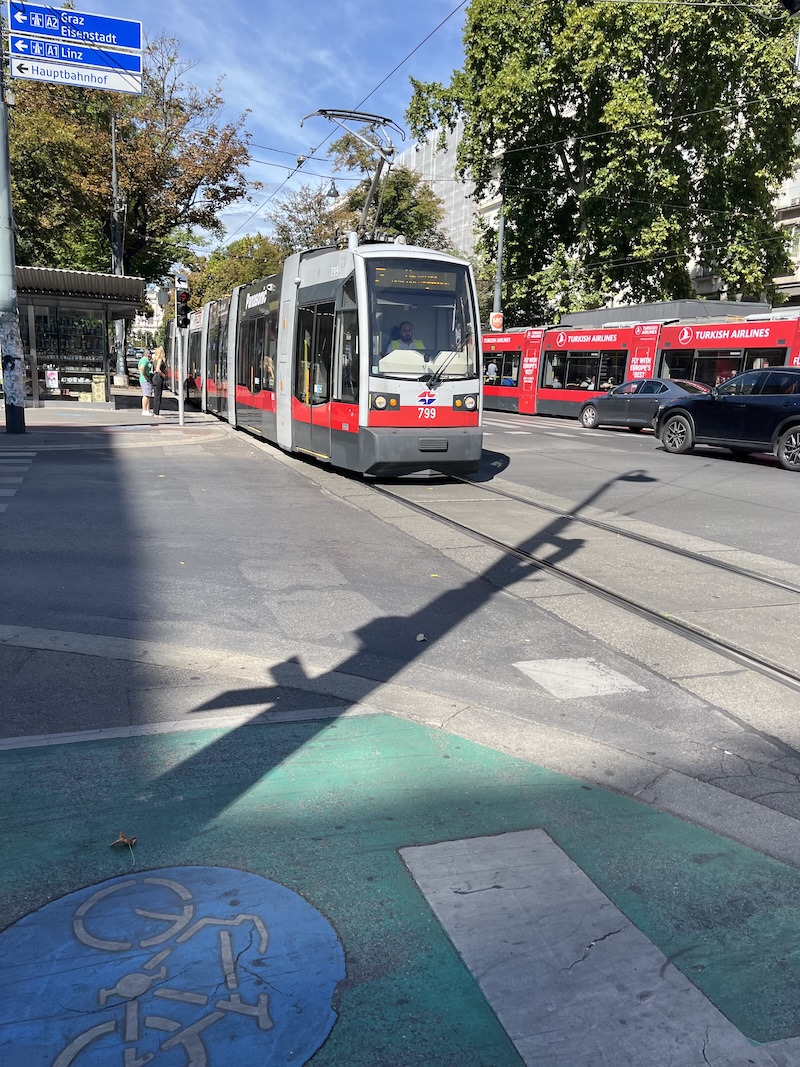 A photo of two trams in Vienna. Each is mainly red. There is also a black car and a grey car in shot, plus a cycle symbol on the tarmac bottom left.