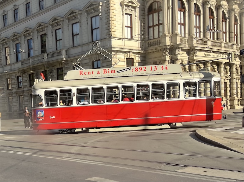 A single red tram car with a cream top displaying 'Rent a Bim - 892 13 34', set against a large building with columns to the right